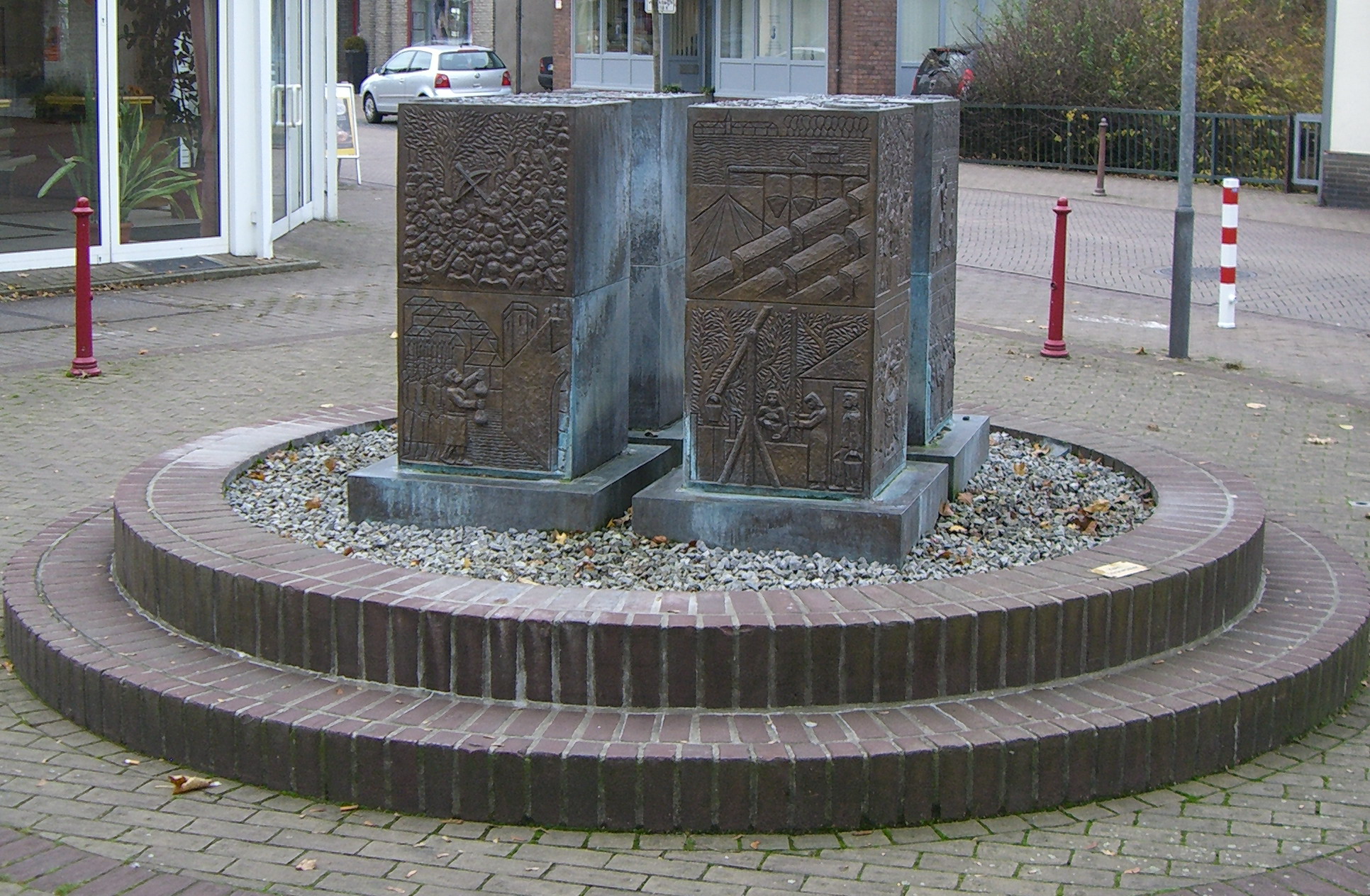 Brunnen bestehend aus 4 rechteckigen Bronzesteelen auf rundgemauertem Sockel